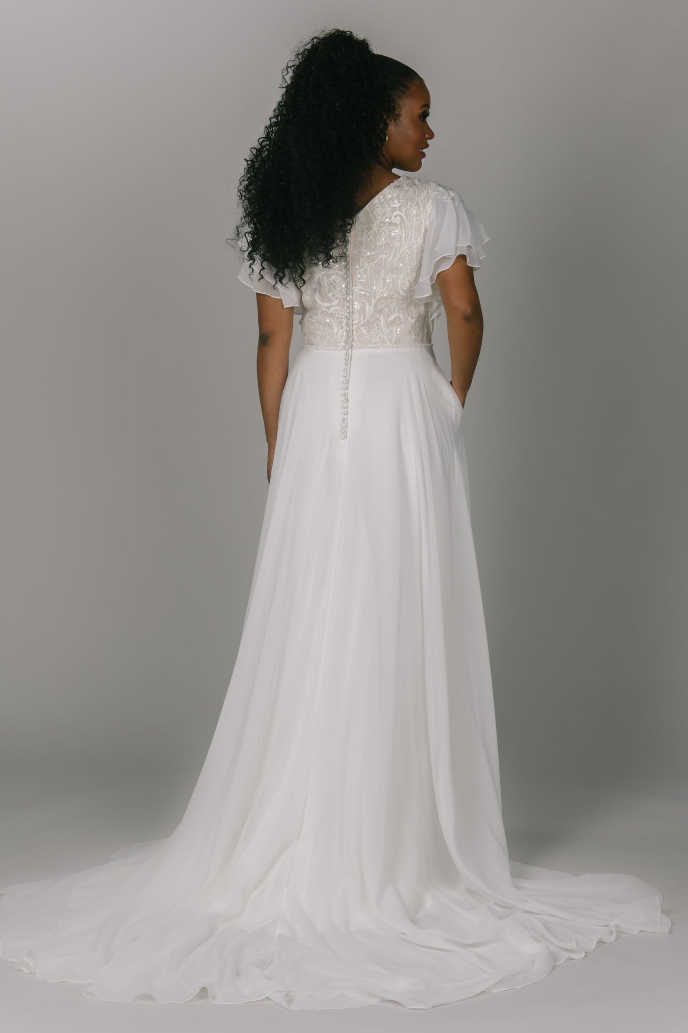 Modest wedding dress with flutter sleeves. It has a v-neckline and an a-line shape. The bottom is a flowy chiffon with a slit. The top is beaded lace. It has covered buttons down the back.