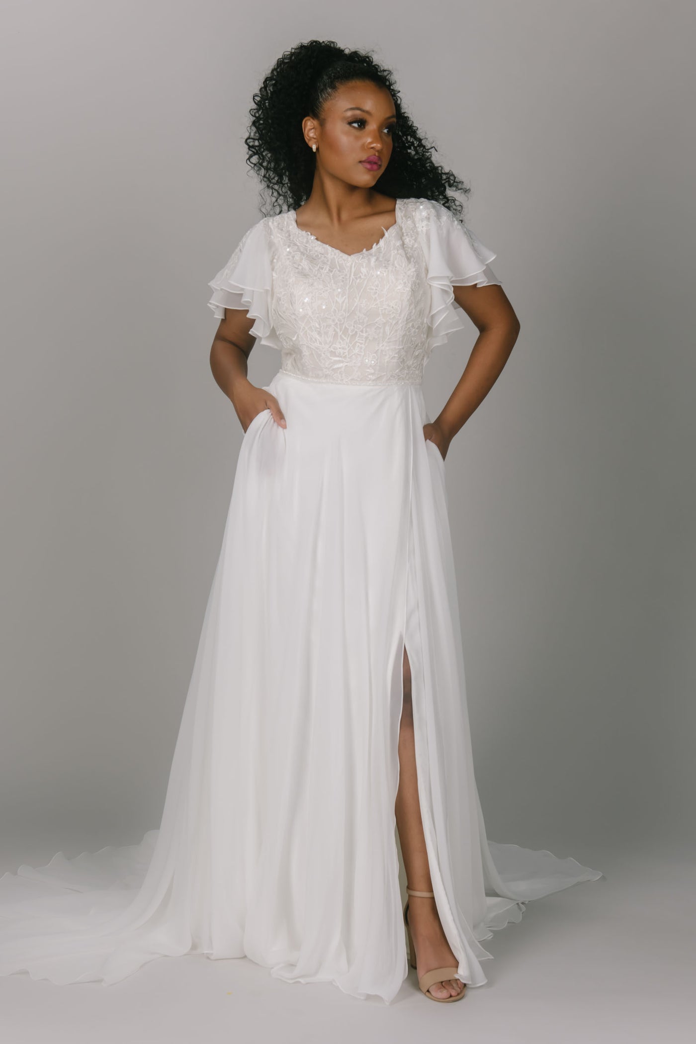 Modest wedding dress with flutter sleeves. It has a v-neckline and an a-line shape. The bottom is a flowy chiffon with a slit. The top is beaded lace. It has covered buttons down the back.