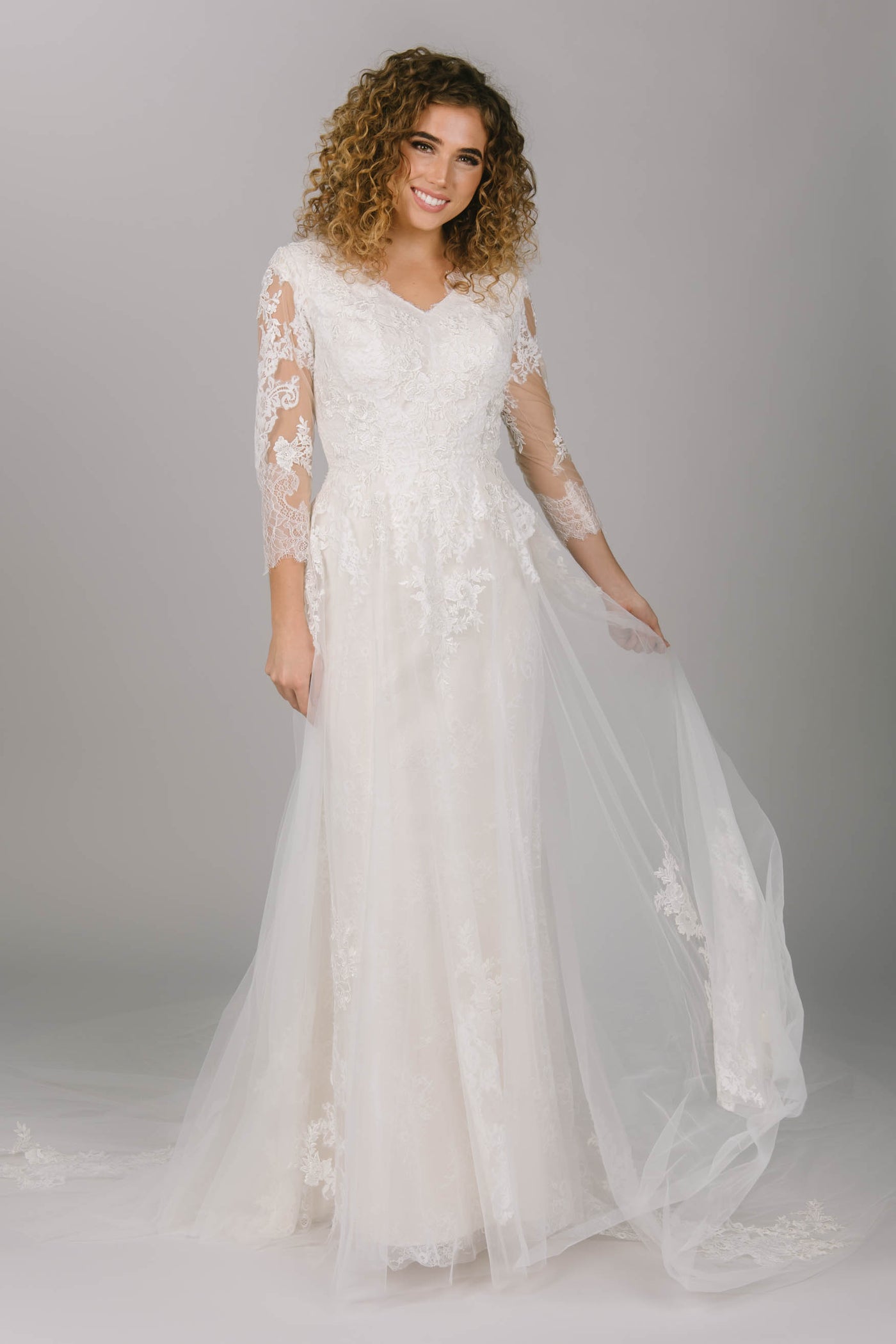 Front view of modest wedding dress with long sleeves. It has a v-neckline and long tulle train. The lace is soft and delicate and is found through the dress. This is a dreamy modest wedding gown.