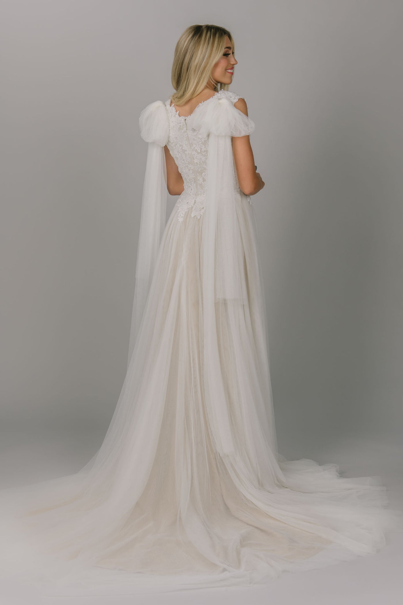 Back view of a-line modest wedding dress. It has a scoop neckline and cap sleeves. The top has lace and beaded detailing. The skirt is tulle with a champagne underlay. This view shows off the tulle bows that come as attachments. 