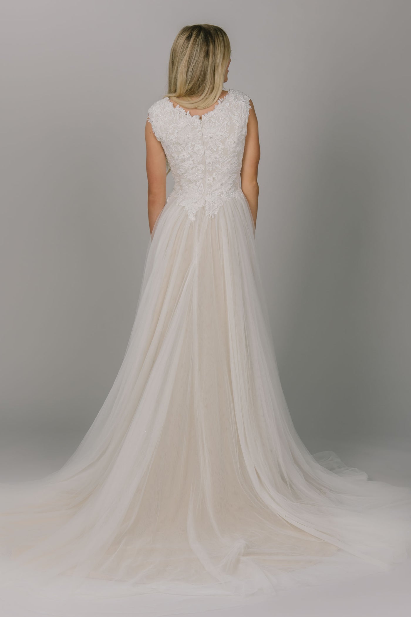 A-line modest wedding dress. It has a scoop neckline and cap sleeves. The top has lace and beaded detailing. The skirt is tulle with a champagne underlay.