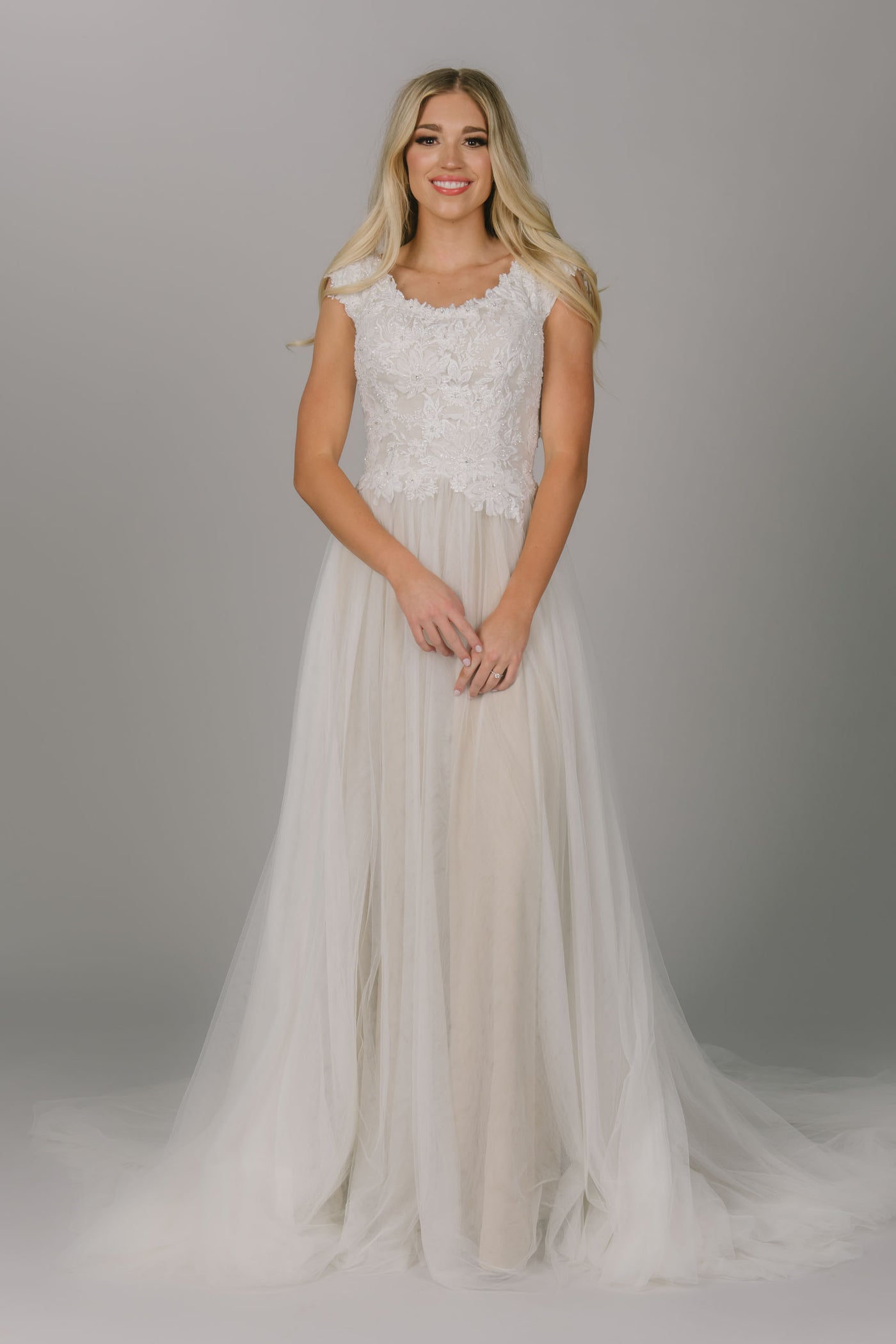 A-line modest wedding dress. It has a scoop neckline and cap sleeves. The top has lace and beaded detailing.  The skirt is tulle with a champagne underlay.  