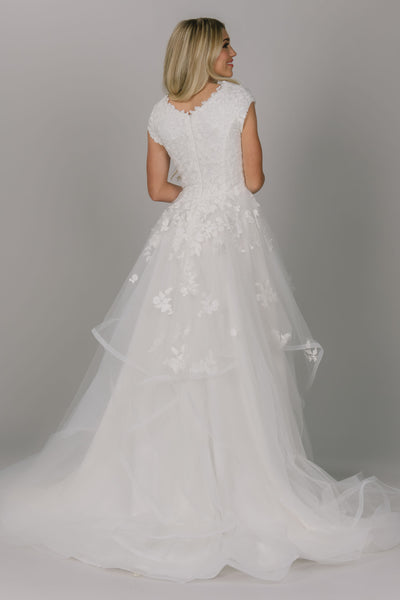 Dreamy modest wedding dress with whimsical skirt. It has flower lace on the top and some on the skirt. It has a scoop neckline and cap sleeves.