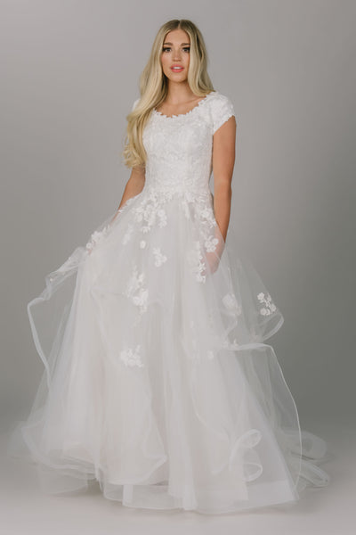 Dreamy modest wedding dress with whimsical skirt. It has flower lace on the top and some on the skirt. It has a scoop neckline and cap sleeves. 