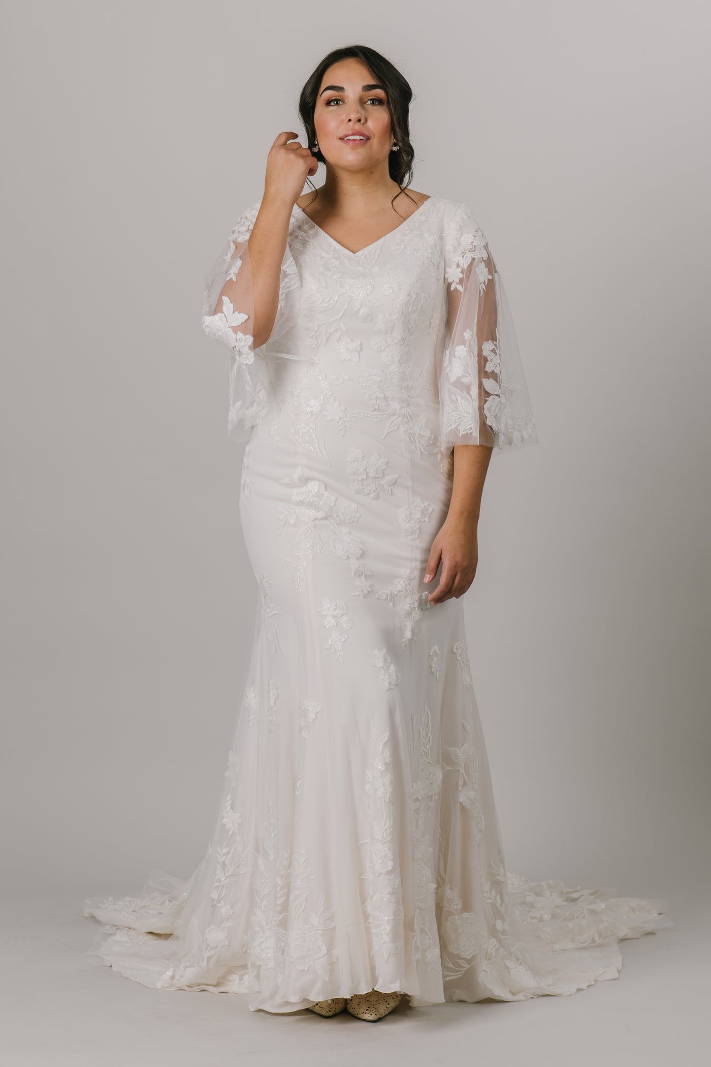 This unique modest wedding gown is like nothing you've seen! It features a v-neckline, fitted silhouette, and gorgeous flowery lace. This gown provides such an amazing fit with details you'll love! Available in Ivory and Ivory/DecoGold.  Style Love: This gown has the most unique flutter sleeves! They're unlike anything else we have in the shop!