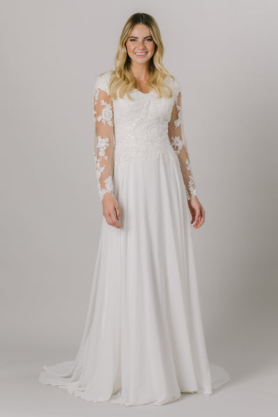 This a-line modest wedding dress is the perfect combination of lace and sparkle. It has a lovely v-neck and illusion long sleeves with buttons up the cuff.  Shown in Ivory/Nude.
