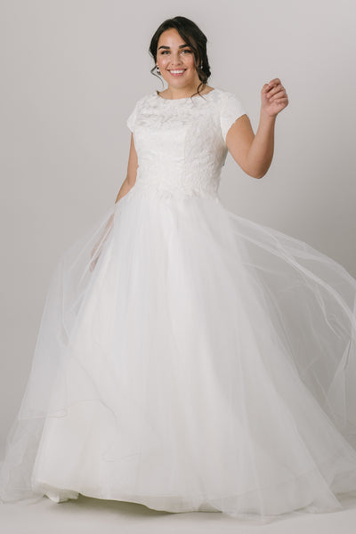 This elegant modest wedding dress is perfect for the traditional bride! We love the soft petal sleeves, the wide boat neckline, and the flattering cinched waist. The gown is shown in Ivory.  Style Love: Add a simple belt to add more sparkle to this gown or keep it simple as is! Feeling like a princess? Add a tiara to complete the look!
