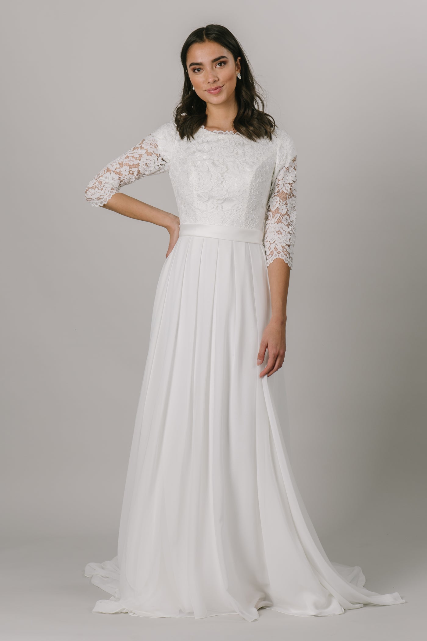 A lace a line white wedding dress that has a belt with a chiffon skirt and buttons that go 3/4 of the dress, from a bridal store located in Salt Lake City, Utah.
