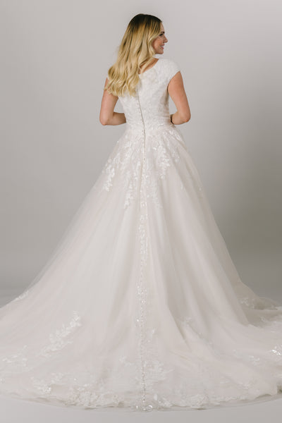 This modest ballgown wedding dress features a flattering fit and gorgeous sparkly floral lace throughout the bodice and trickling down the skirt. It has a soft v-neck and buttons down the back! Available in Ivory and Ivory/Champagne (pictured).  Style Love: Appliques from the top and bottom meet at the middle of the skirt! From a bridal shop in downtown Salt Lake City.