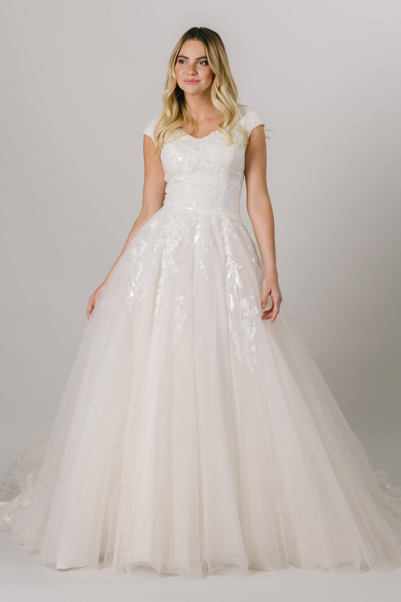 This modest ballgown wedding dress features a flattering fit and gorgeous sparkly floral lace throughout the bodice and trickling down the skirt. It has a soft v-neck and buttons down the back! Available in Ivory and Ivory/Champagne (pictured).  Style Love: Appliques from the top and bottom meet at the middle of the skirt!