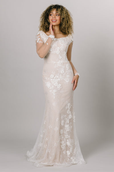 This modest wedding gown is complete with a fitted silhouette, long mesh sleeves, stunning 3D applique flowers, and perfected with a delicate lace underlay. 