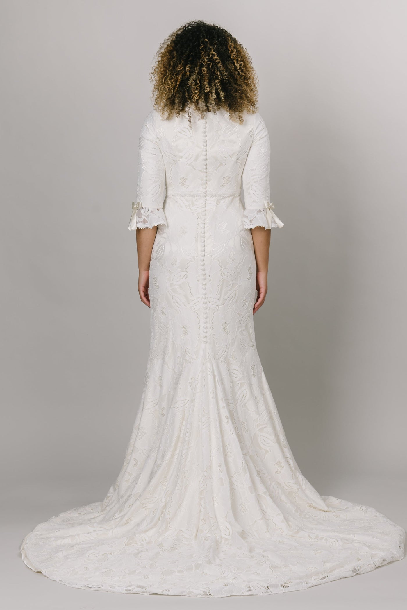 The back view of a modest wedding dress features all-over floral lace, in a fitted silhouette. The neckline has a dainty trim that matches the cute bell sleeve!