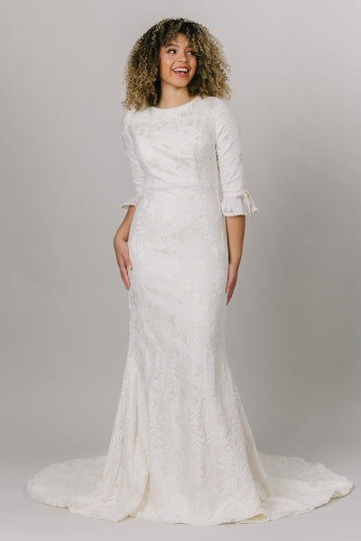 A modest wedding dress features all-over floral lace, in a fitted silhouette. The neckline has a dainty trim that matches the cute bell sleeve!