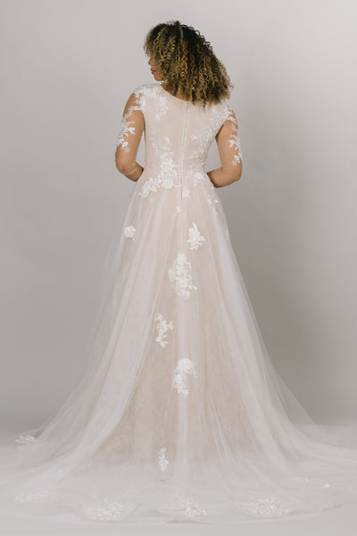 A back view of the modest wedding dress is the whimsical gown of our dreams! With a soft lace print under the top layer of appliques, mesh sleeves, and a scoop neck, you'll be dancing the night away. 