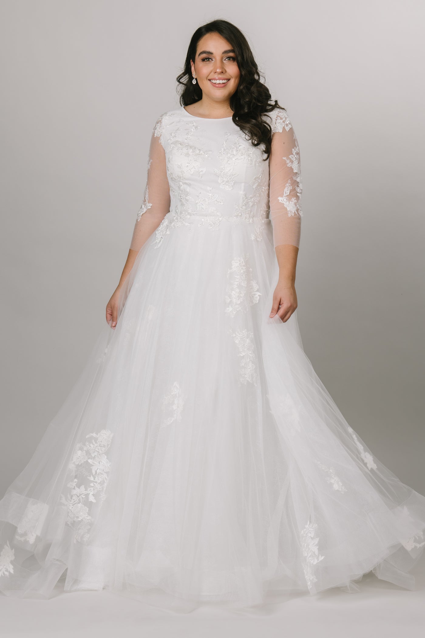 In a white color this modest wedding dress is the whimsical gown of our dreams! With a soft lace print under the top layer of appliques, mesh sleeves, and a scoop neck, you'll be dancing the night away. 