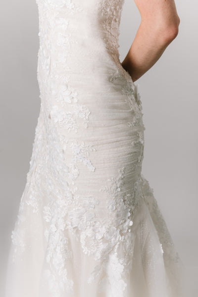 Close up view of a modest wedding dress with a fitted silhouette, cap sleeve, and v-neckline. The lace drapes down the skirt of the dress. 