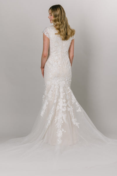 Back view of a modest wedding dress with a fitted silhouette, cap sleeve, and v-neckline. The lace drapes down the skirt of the dress. 