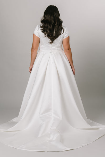 Back view of this modest wedding dress with cap sleeves and v-neckline. This sleek an elegant Moments Made dress has pockets. This dress has comes with a thin and simple belt. It has a zipper back and pleats to create a beautiful flowing a-line cut.