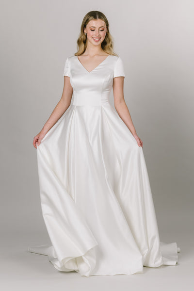 Front view of this modest wedding dress with cap sleeves and v-neckline. This sleek an elegant Moments Made dress has pockets. This dress has comes with a thin and simple belt. It has a zipper back and pleats to create a beautiful flowing a-line cut.