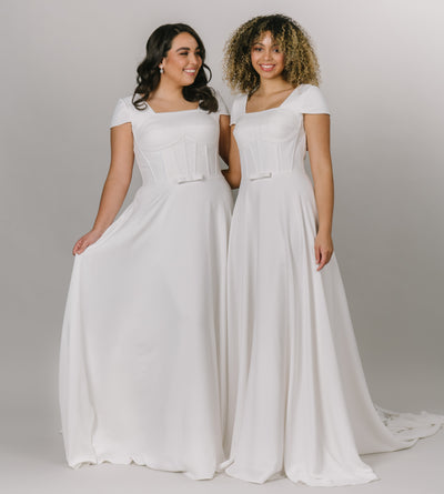 Two models both wearing the same model wearing modest wedding dress that is a-line with a square neckline. It has a more constructed top with cap sleeves.