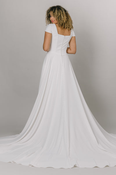 Back view of model wearing modest wedding dress that is a-line with a square neckline. It has a more constructed top with cap sleeves.