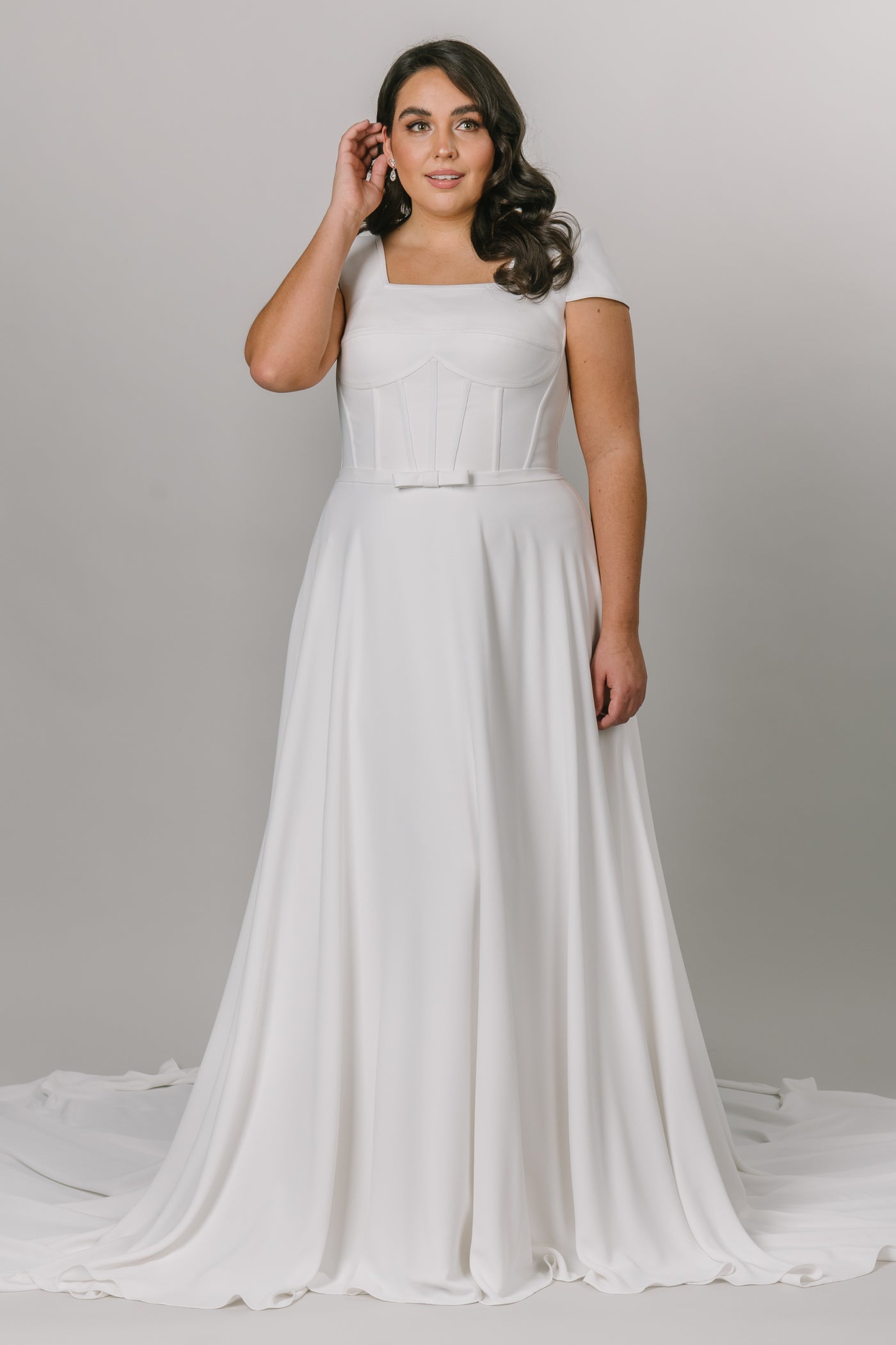 Plus size model wearing modest wedding dress that is a-line with a square neckline. It has a more constructed top with cap sleeves. 