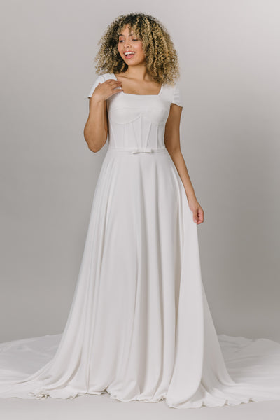 Front view of model wearing modest wedding dress that is a-line with a square neckline. It has a more constructed top with cap sleeves.