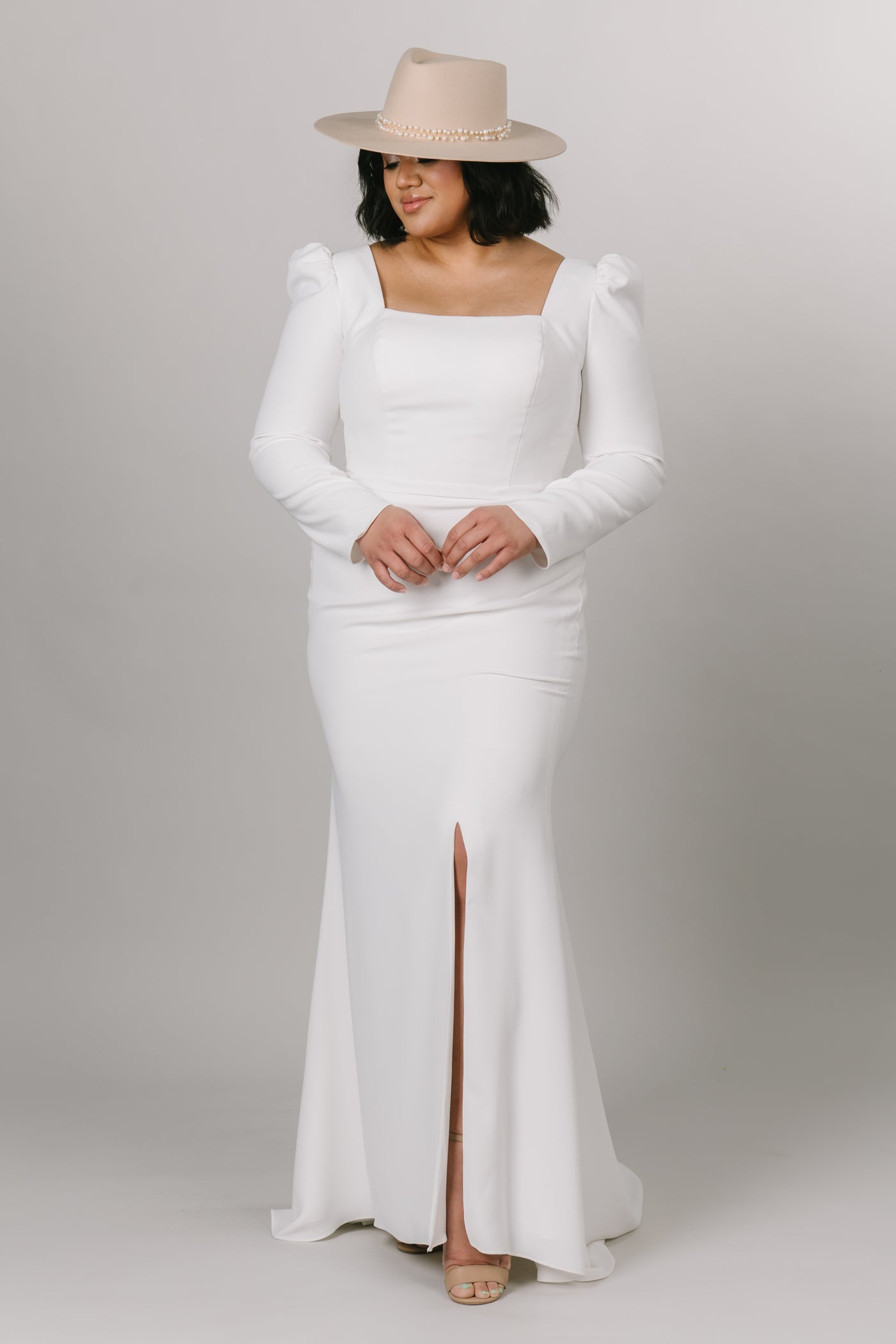 Front view of modest wedding dress with long puffed sleeves. This dress is fitted with a knee high slit and a square neck line. Model is styled with a boho hat to create the overall look.