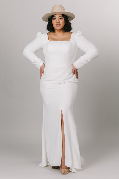 Front view of modest wedding dress with long puffed sleeves. This dress is fitted with a knee high slit and a square neck line. Model is styled with a boho hat to create the overall look. 