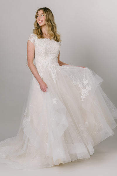 Modest wedding dress with square neckline and a-line fit. It has cap sleeves and tulip tiered skirt. It has lace on the bodice and on the edges of the tiered skirt. 