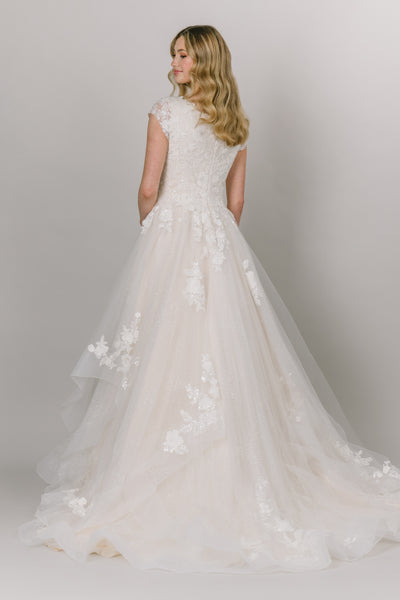 Back view of modest wedding dress with square neckline and a-line fit. It has cap sleeves and tulip tiered skirt. It has lace on the bodice and on the edges of the tiered skirt.