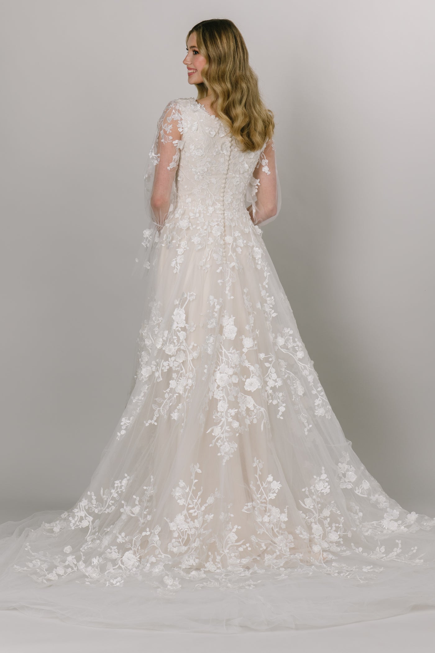 Back view of gorgeous modest wedding dress with a-line fit and heart shaped neckline. It has trumpet-link tulle sleeves with delicate flower lace. The flower lace is on the bodice and tiered towards the bottom.
