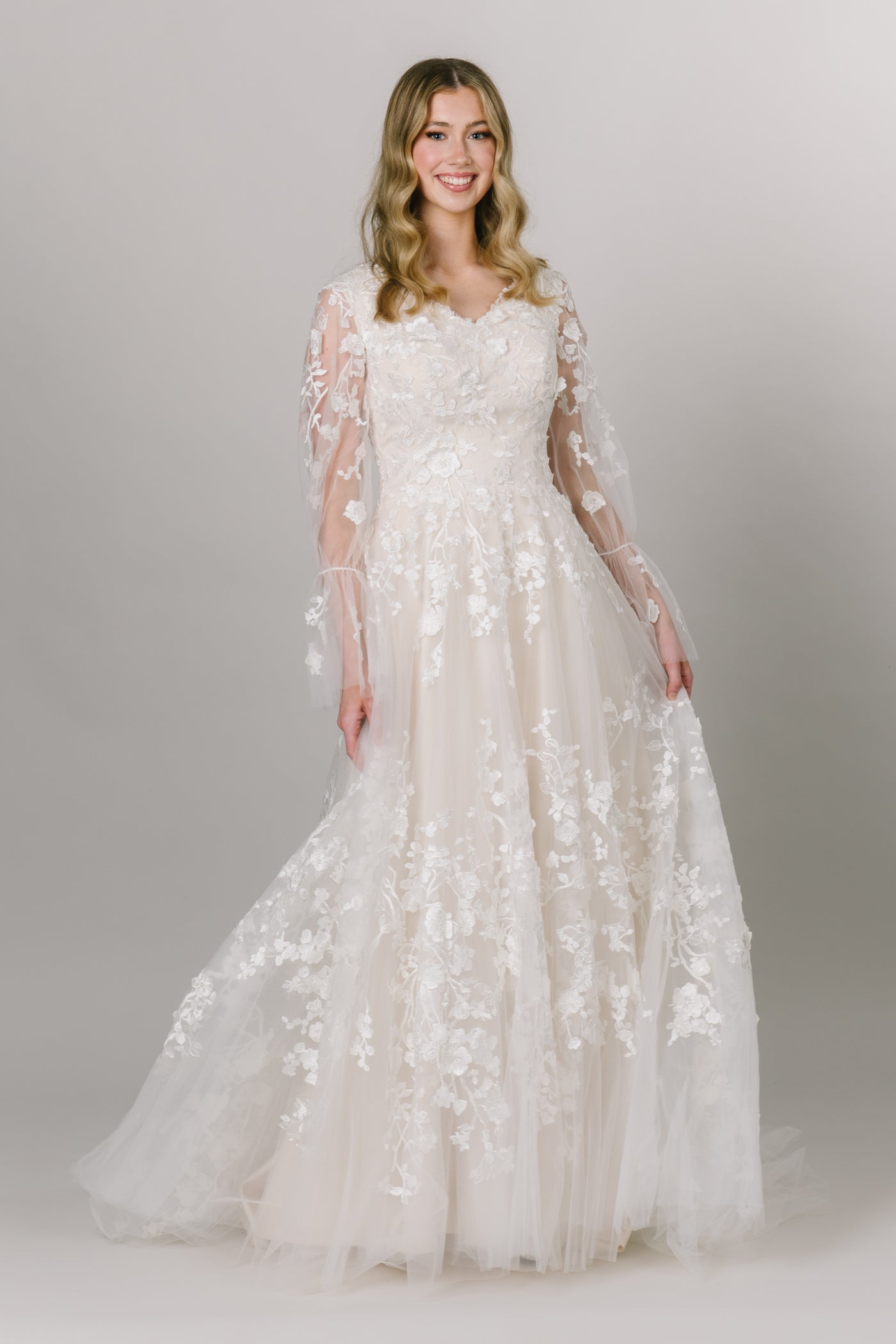 Front view of gorgeous modest wedding dress with a-line fit and heart shaped neckline. It has trumpet-link tulle sleeves with delicate flower lace. The flower lace is on the bodice and tiered towards the bottom. 