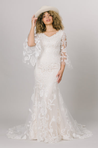 This modest wedding dress has a lace pattern and complemented with beautiful bell sleeves!, featuring a cream hat.