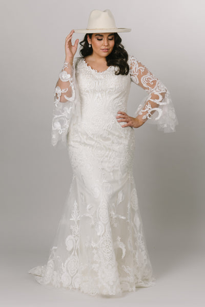 Front view of a modest wedding dress features a stunning lace pattern over a fitted silhouette with a cream hat.