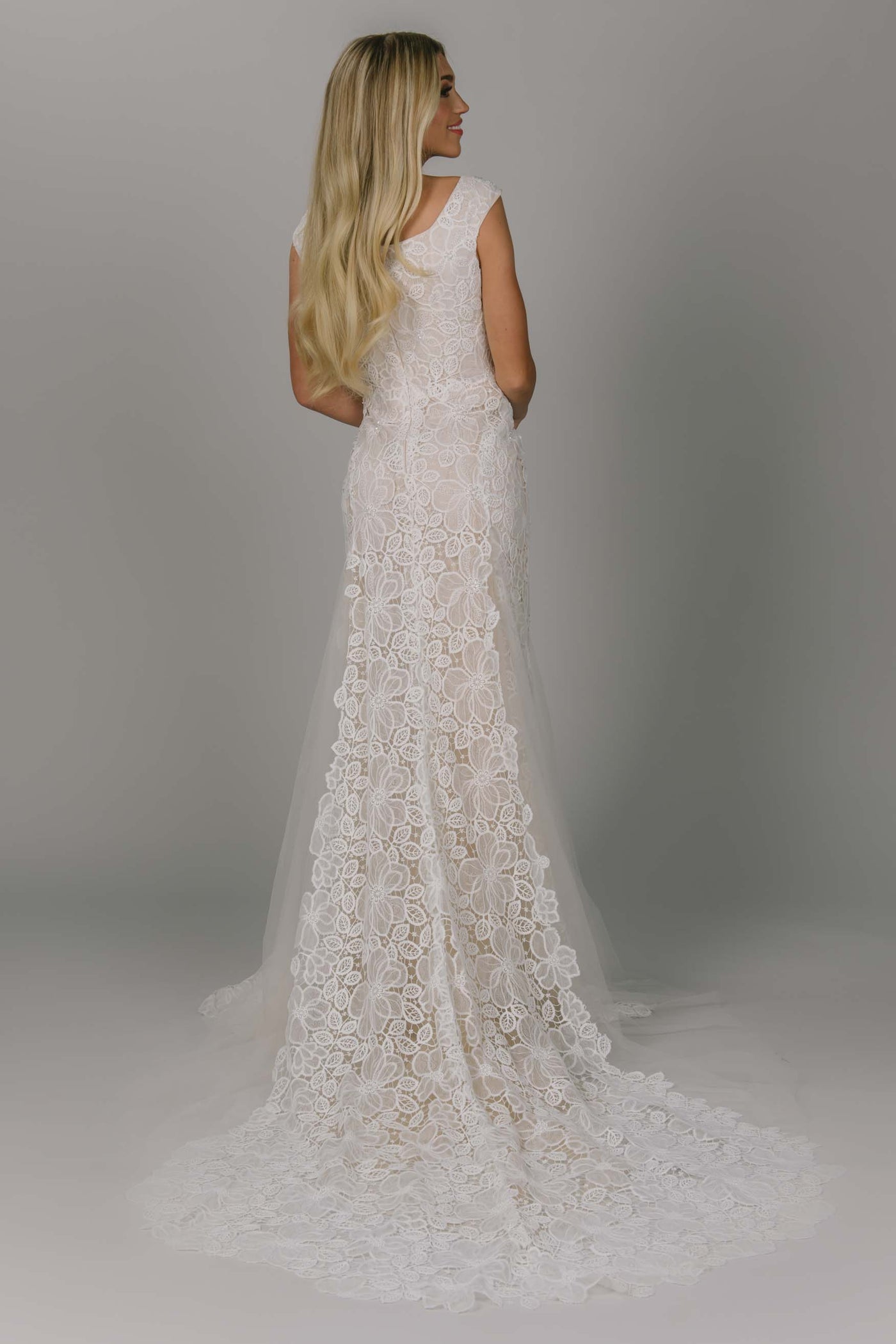 Fitted modest wedding dress featuring flower petal lace. It has a v-neckline and champagne underlay. It has cap sleeves and tulle slits. Absolutely stunning for any modest wedding dress bride.