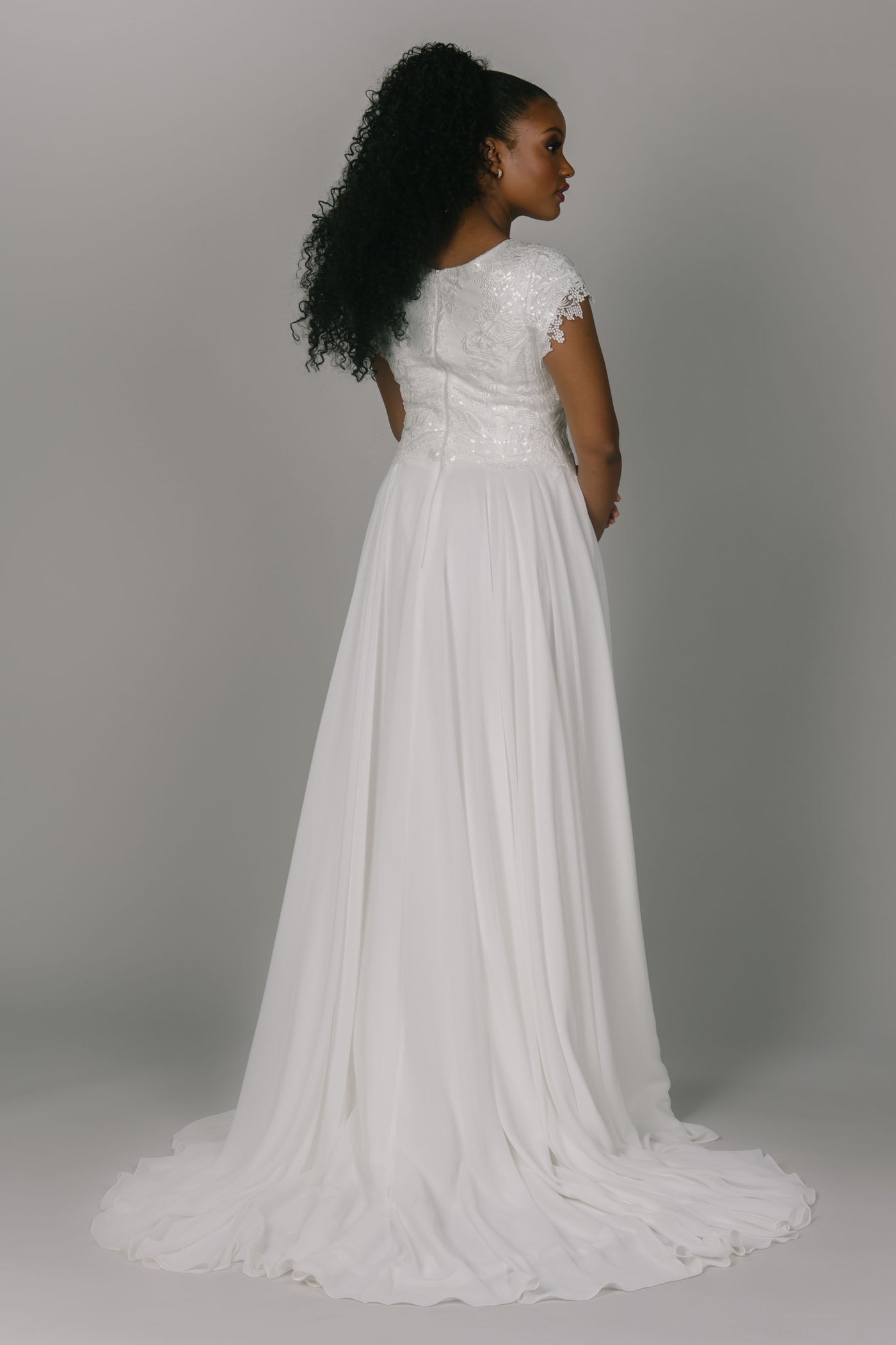 Modest wedding dress featuring an a-line cut. It has a lace and beaded top with a chiffon bottom. It has cap sleeves and a zipper back. This dress is perfect for the modest wedding dress bride looking for something between a a-line and fitted wedding dress.