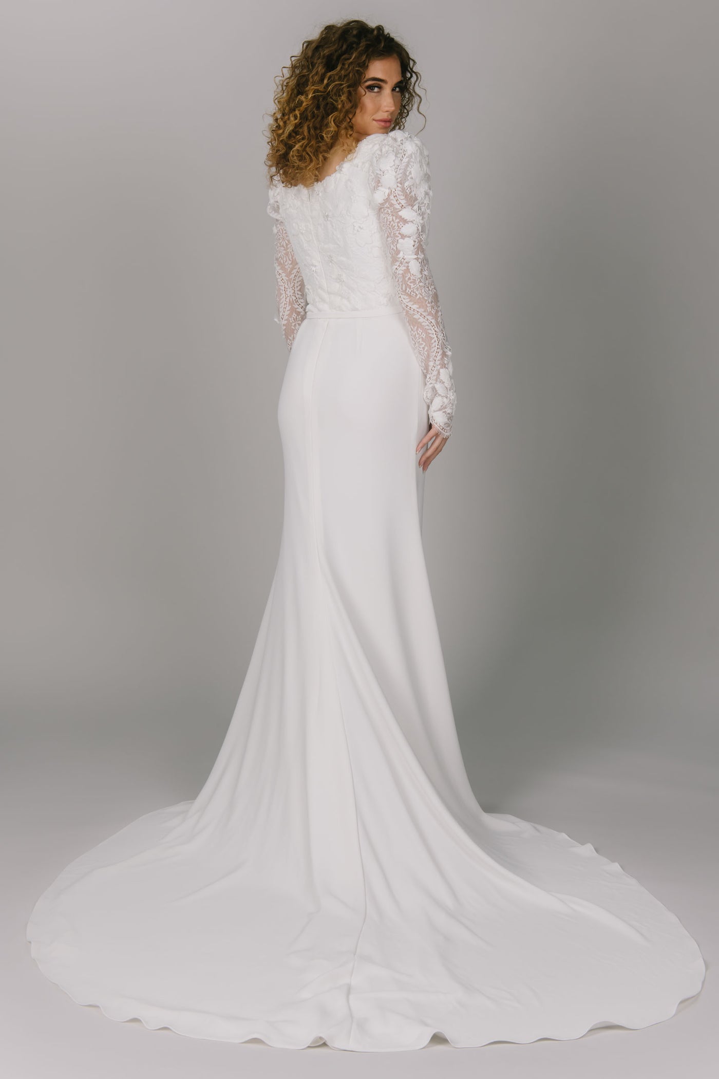 Back view of modest wedding dress with sleeves. It has lace puff sleeves and a sweetheart neckline. It has a simple skirt with a slit. This modest wedding gown comes with a bow belt.