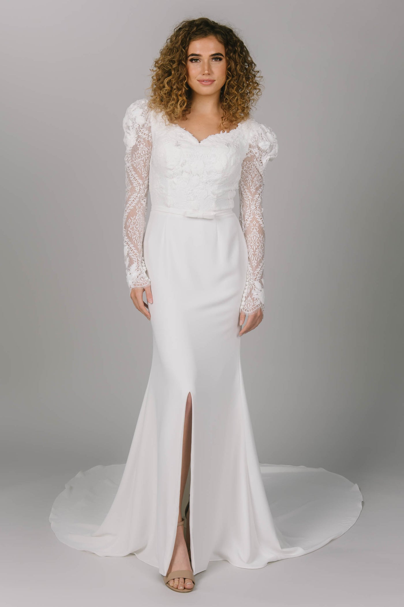 Front view of modest wedding dress with sleeves. It has lace puff sleeves and a sweetheart neckline. It has a simple skirt with a slit. This modest wedding gown comes with a bow belt. 