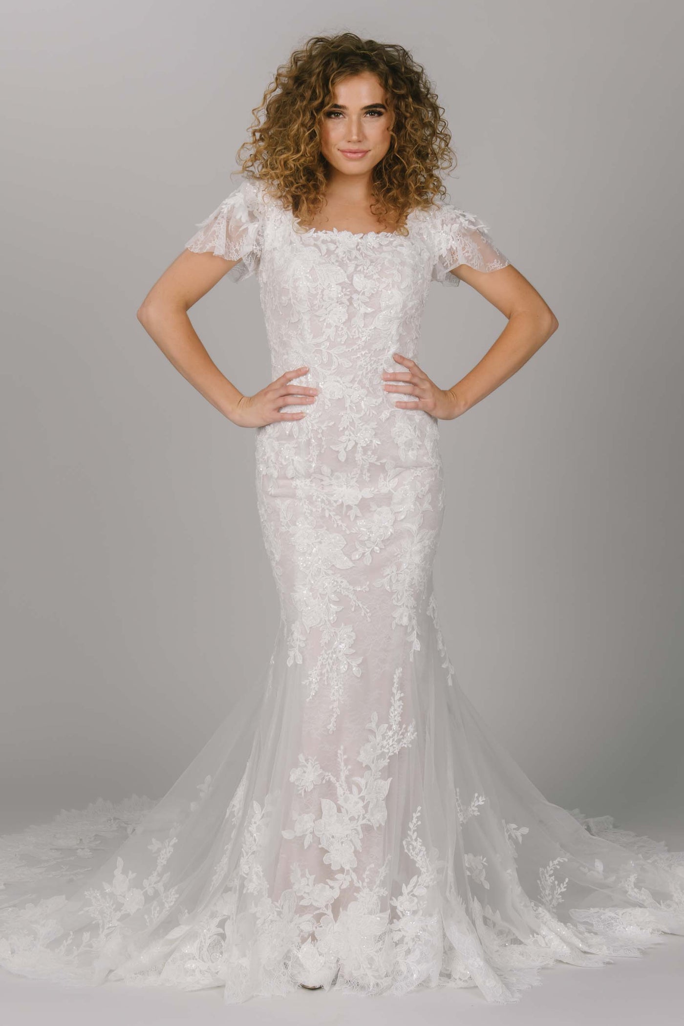 Front view of modest wedding with lace and beading. This dress has a fitted silhouette with a long train. It has a square neckline. This modest wedding dress is perfect for the show-stopping bride.  